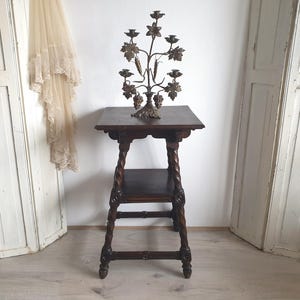 May include: A dark brown wooden side table with a lower shelf and carved legs. A decorative metal candelabra with leaf and grape details sits on top. The candelabra has space for multiple candles.