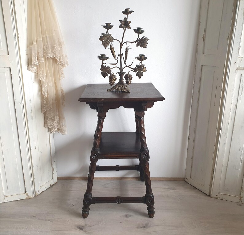 May include: A dark brown wooden side table with a lower shelf and carved legs. A decorative metal candelabra with leaf and grape details sits on top. The candelabra has space for multiple candles.