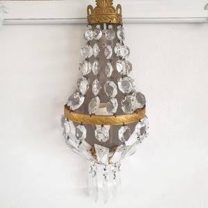 Puede incluir: Aplique de pared antiguo con marco de metal dorado y detalles de cristal transparente. El aplique presenta una parte superior decorativa, una secci&oacute;n central con cristales colgantes y una secci&oacute;n inferior con colgantes de cristal adicionales. Montado en una pared blanca.