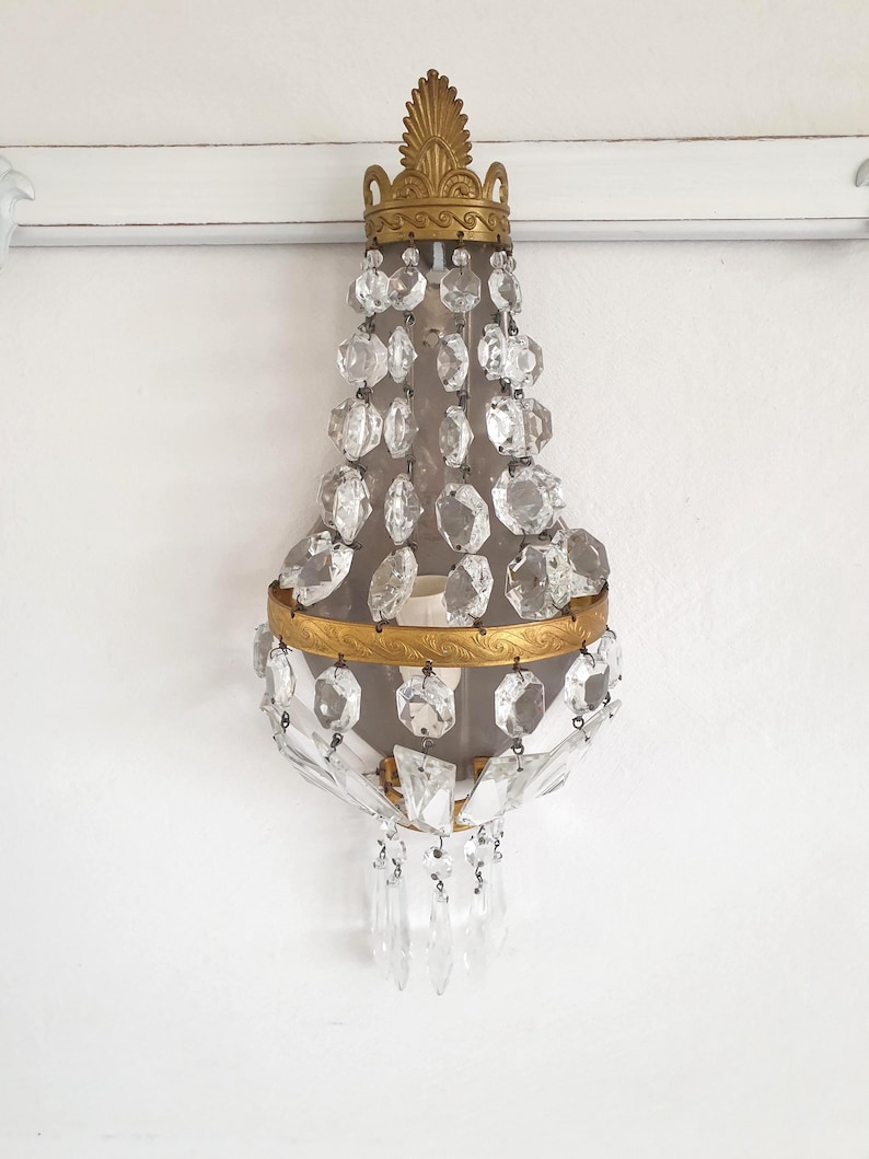 Puede incluir: Aplique de pared antiguo con marco de metal dorado y detalles de cristal transparente. El aplique presenta una parte superior decorativa, una secci&oacute;n central con cristales colgantes y una secci&oacute;n inferior con colgantes de cristal adicionales. Montado en una pared blanca.