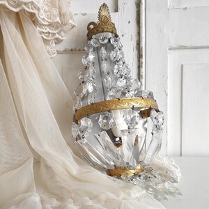 Puede incluir: Una l&aacute;mpara de ara&ntilde;a de cristal vintage con un marco de metal dorado y detalles decorativos. La l&aacute;mpara est&aacute; adornada con numerosos colgantes y cuentas de cristal transparente. El fondo incluye una puerta blanca y una cortina de encaje color crema.