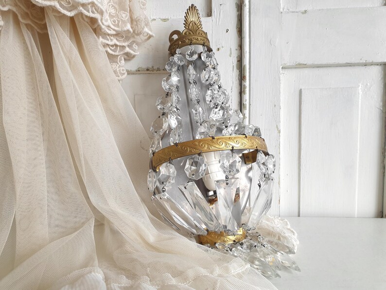 Puede incluir: Una l&aacute;mpara de ara&ntilde;a de cristal vintage con un marco de metal dorado y detalles decorativos. La l&aacute;mpara est&aacute; adornada con numerosos colgantes y cuentas de cristal transparente. El fondo incluye una puerta blanca y una cortina de encaje color crema.