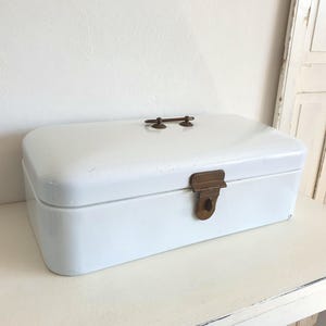 Könnte beinhalten: Weiße Email-Brotdose mit einem Messingverschluss und zwei Messinggriffen. Die Brotdose steht auf einem weißen Tisch.