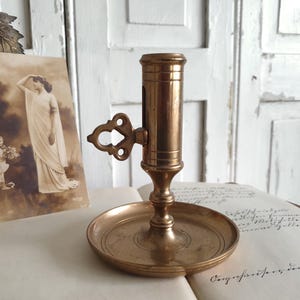 May include: A vintage brass candlestick holder with a round base and a cylindrical top. The holder features an ornate handle and is set on an open book with a sepia-toned postcard in the background. The candlestick is a warm, antique gold color.