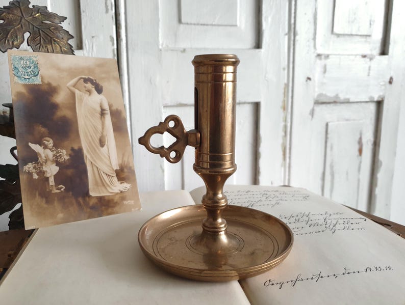 May include: A vintage brass candlestick holder with a round base and a cylindrical top. The holder features an ornate handle and is set on an open book with a sepia-toned postcard in the background. The candlestick is a warm, antique gold color.