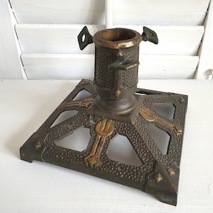 Può includere: Un porta albero di Natale vintage in ghisa con una base quadrata e una colonna centrale. Il supporto è decorato con un motivo geometrico e ha una finitura dorata. Il supporto ha quattro bracci che si estendono dalla base e sono progettati per sostenere un albero di Natale.