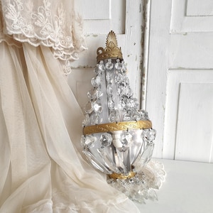 Puede incluir: Una l&aacute;mpara de pared de estilo vintage con detalles de cristal transparente y adornos de metal dorado. La l&aacute;mpara tiene una parte superior e inferior decorativas y est&aacute; rodeada de tela transparente de color crema.