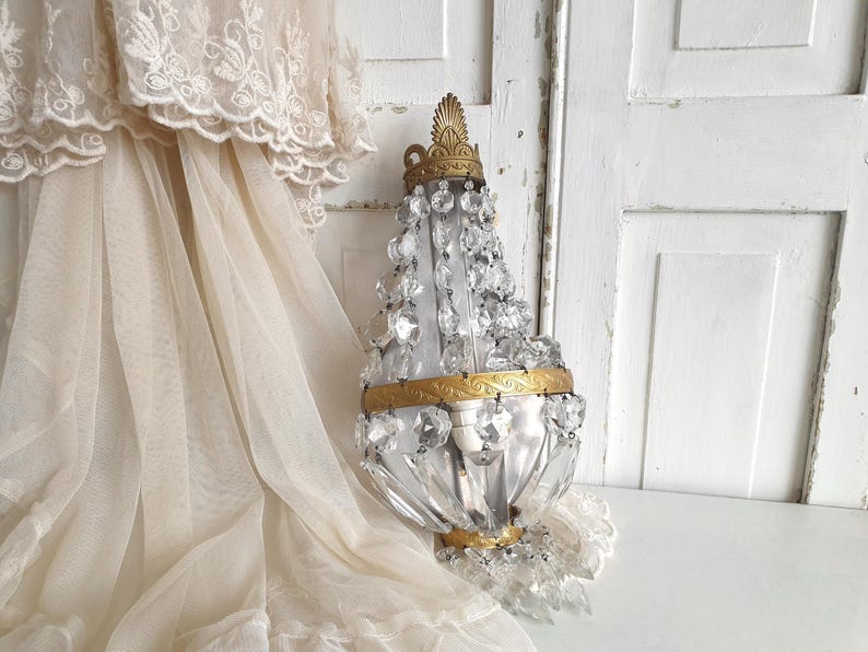 Puede incluir: Una l&aacute;mpara de pared de estilo vintage con detalles de cristal transparente y adornos de metal dorado. La l&aacute;mpara tiene una parte superior e inferior decorativas y est&aacute; rodeada de tela transparente de color crema.