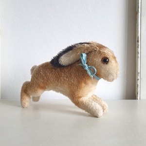Può includere: Un giocattolo vintage a forma di coniglio di peluche in posizione di corsa. Il coniglio ha la pelliccia marrone con una striscia nera sulla schiena e un nastro azzurro intorno al collo. Il giocattolo è ambientato su uno sfondo bianco.