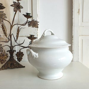 Puede incluir: Una fuente blanca con tapa, cuerpo redondeado y dos asas pequeñas. La fuente es de cerámica y está sobre una superficie blanca. Un candelabro decorativo de bronce con detalles de uvas y hojas está en el fondo.