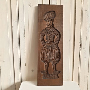 Puede incluir: Un molde de galletas antiguo de madera rectangular con una figura tallada de una persona con vestimenta tradicional. La figura lleva un sombrero, una camisa abotonada y pantalones abombados. El molde es de color marr&oacute;n c&aacute;lido.