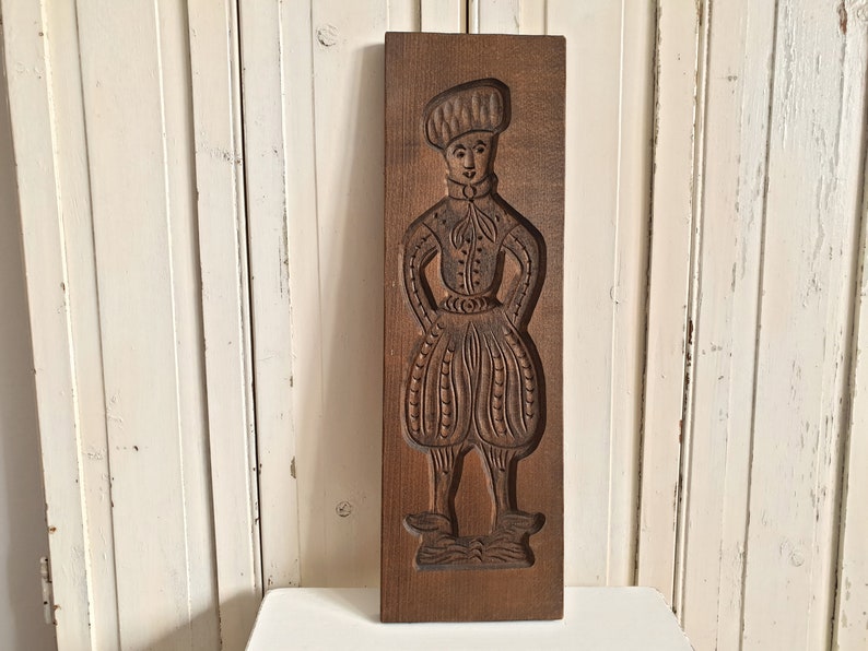 Puede incluir: Un molde de galletas antiguo de madera rectangular con una figura tallada de una persona con vestimenta tradicional. La figura lleva un sombrero, una camisa abotonada y pantalones abombados. El molde es de color marr&oacute;n c&aacute;lido.