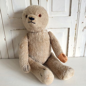 May include: A vintage beige teddy bear with black eyes and a stitched nose. The bear has brown felt patches on its paws and is sitting on a white surface.