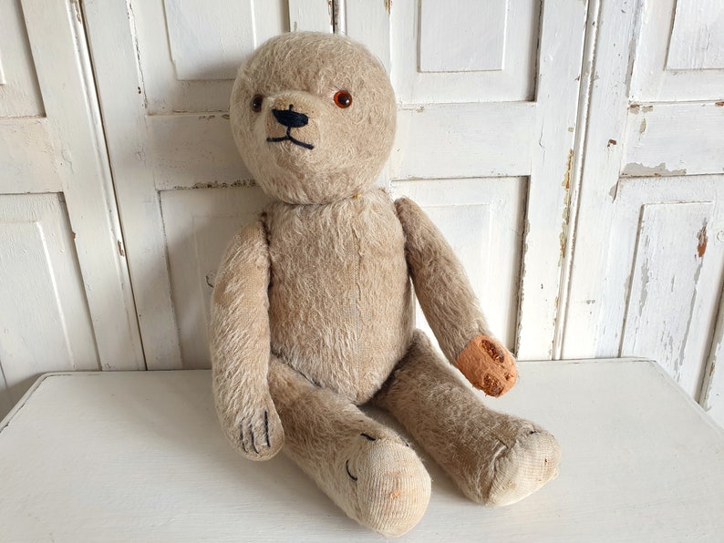 May include: A vintage beige teddy bear with black eyes and a stitched nose. The bear has brown felt patches on its paws and is sitting on a white surface.