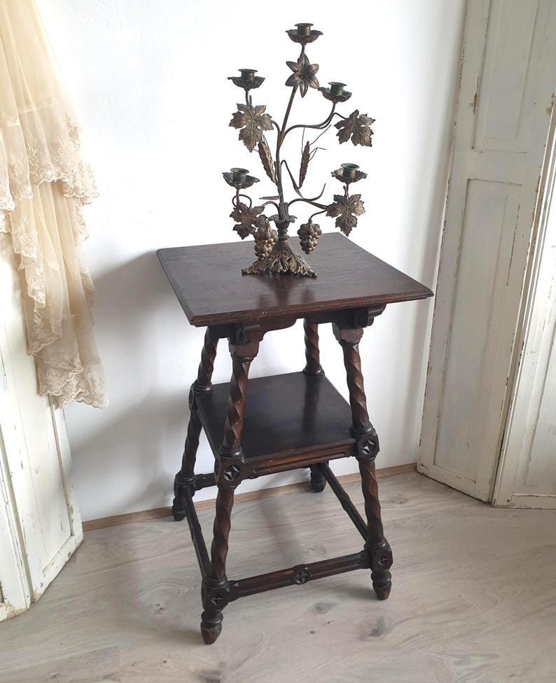 May include: A dark brown, antique wooden side table with a lower shelf and intricately carved legs. A decorative brass candelabra with leaf and grape details sits on top. The candelabra has multiple candle holders.