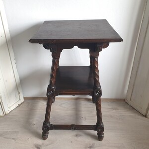 May include: Dark brown antique side table with a square top and a lower shelf. The table features intricately carved details and twisted legs. The table is approximately 28 inches tall.