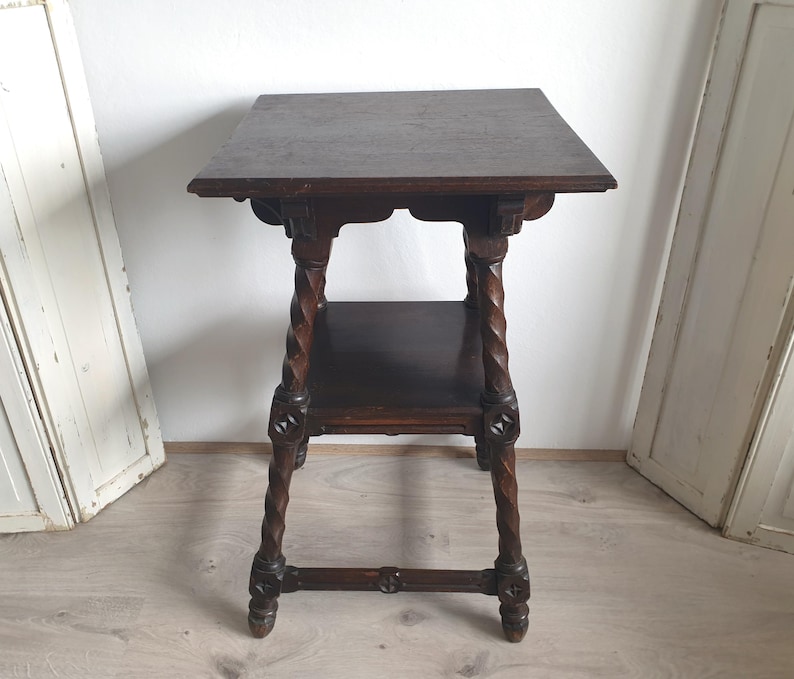 May include: Dark brown antique side table with a square top and a lower shelf. The table features intricately carved details and twisted legs. The table is approximately 28 inches tall.