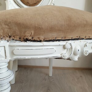 Puede incluir: Silla de madera pintada de blanco con un asiento de tela marr&oacute;n desgastado. La silla tiene un dise&ntilde;o tallado en el respaldo y las patas.
