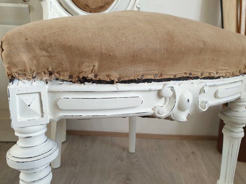 Puede incluir: Silla de madera pintada de blanco con un asiento de tela marr&oacute;n desgastado. La silla tiene un dise&ntilde;o tallado en el respaldo y las patas.