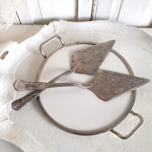 Può includere: Un set da portata per torte argentato su un vassoio da portata bianco e argento. Il set include due palette per torte con disegni ornati e un vassoio rotondo con manici. Il vassoio ha un centro bianco e un bordo argentato.