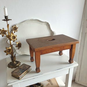 Könnte beinhalten: Ein rechteckiger Holzhocker mit vier stabilen Beinen und einer ovalen Grifföffnung in der Mitte, neben einem goldfarbenen Metallkerzenhalter in Form eines Blumenstraußes und einer kleinen rechteckigen Metallschachtel mit Verzierungen.