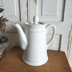 May include: A white ceramic coffee pot with a lid and a curved spout. The pot has a handle and sits on a wooden surface. The background features a white door and a decorative candle holder.
