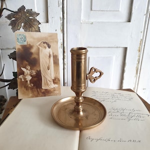 May include: A vintage brass candlestick holder with a round base and a handle with a decorative cutout. An antique postcard with a sepia-toned image of a woman and an angel is next to the candlestick. The items are on a book with handwritten text.