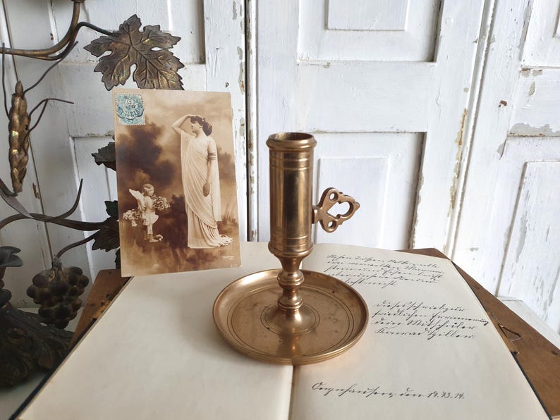 May include: A vintage brass candlestick holder with a round base and a handle with a decorative cutout. An antique postcard with a sepia-toned image of a woman and an angel is next to the candlestick. The items are on a book with handwritten text.