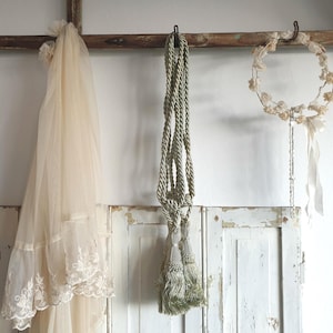 May include: A collection of vintage-style decorative items hanging against a white wall. Includes a cream-colored veil with lace trim, a twisted rope with tassels, and a floral wreath with ribbons. The items are suspended from a wooden beam.
