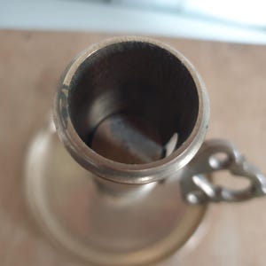 May include: A vintage brass candlestick holder with a circular base and a handle with a decorative design. The interior of the candle holder is dark, and the exterior has a tarnished appearance.