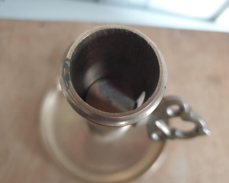 May include: A vintage brass candlestick holder with a circular base and a handle with a decorative design. The interior of the candle holder is dark, and the exterior has a tarnished appearance.