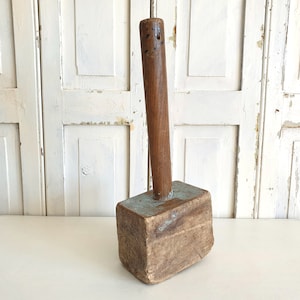 May include: A large wooden mallet with a blue painted head and a brown wooden handle. The mallet is sitting on a white surface.