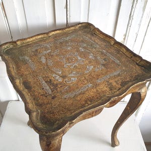 May include: A small, antique, gold-toned table with a scalloped edge. The tabletop features an ornate, textured design. The table has curved legs and is likely made of wood. The table is approximately 24 inches wide.