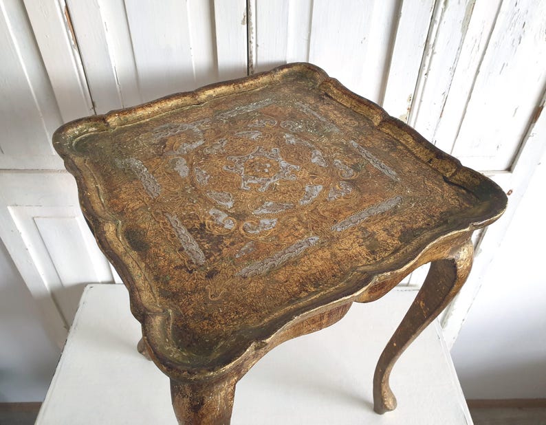 May include: A small, antique, gold-toned table with a scalloped edge. The tabletop features an ornate, textured design. The table has curved legs and is likely made of wood. The table is approximately 24 inches wide.