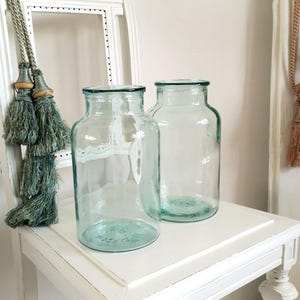 May include: Two clear glass jars with a slight blue-green tint sit on a white table. The jars have a wide, round shape with a narrow neck and a dark rim. Decorative tassels hang in the background.