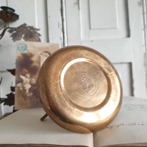 May include: A vintage, round, copper-colored metal object with a small stand. The bottom of the object has the text "Ellis Malat 1997" embossed in a circular pattern. The object is resting on an open book.
