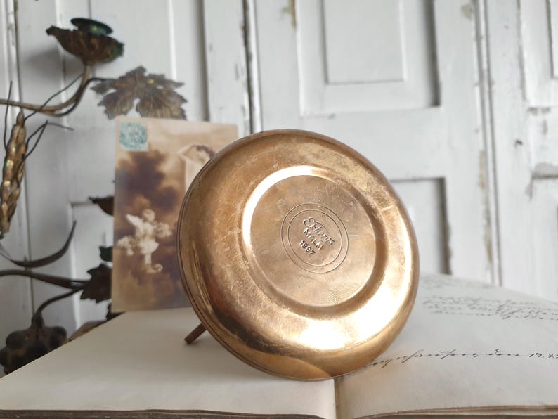 May include: A vintage, round, copper-colored metal object with a small stand. The bottom of the object has the text "Ellis Malat 1997" embossed in a circular pattern. The object is resting on an open book.