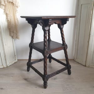May include: A dark brown, antique wooden side table with a square top and a lower shelf. The table features intricately carved details and twisted legs, showcasing a vintage aesthetic. The table is approximately 28 inches tall.