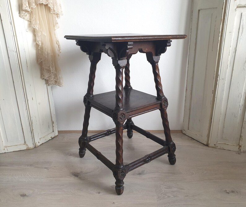 May include: A dark brown, antique wooden side table with a square top and a lower shelf. The table features intricately carved details and twisted legs, showcasing a vintage aesthetic. The table is approximately 28 inches tall.