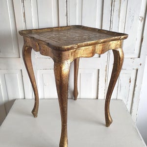 May include: A small, antique gold-colored side table with a square top and curved legs. The table has a distressed finish, giving it a vintage appearance. The table's height is approximately 24 inches.
