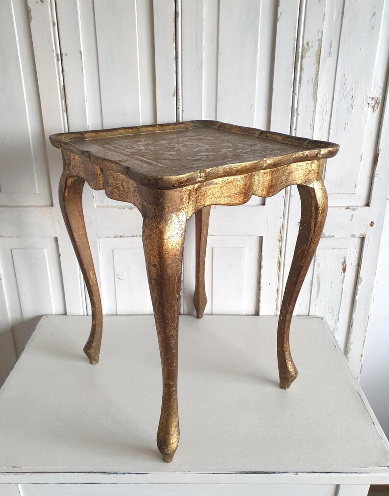 May include: A small, antique gold-colored side table with a square top and curved legs. The table has a distressed finish, giving it a vintage appearance. The table's height is approximately 24 inches.
