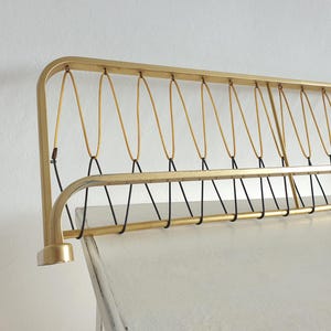 May include: A gold-colored metal wall-mounted coat rack with a rectangular design. The rack features a series of curved lines and black cords, with hooks for hanging items. The design is simple and elegant.