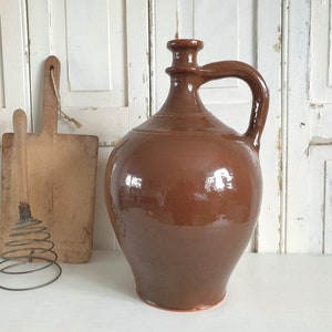 May include: A large, brown, ceramic pitcher with a wide mouth and a handle. The pitcher has a glossy finish and is slightly rounded.