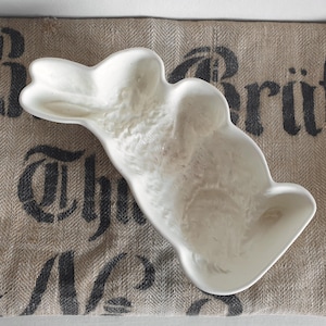Könnte beinhalten: Eine weiße, hasenförmige Keramikschale. Die Schale befindet sich auf einem stoffähnlichen Stoff mit schwarzem Schablonentext. Die Schale hat eine strukturierte Oberfläche und eine flache Schalenform, ideal zum Servieren von Snacks.