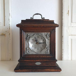 Puede incluir: Reloj de sobremesa antiguo de madera con esfera plateada. La esfera del reloj presenta números romanos y detalles de querubines. El reloj tiene un asa en la parte superior y una pequeña placa decorativa en la base. El reloj mide unos 25 cm de alto.