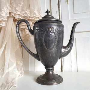 Op de afbeelding: Een vintage zilveren theepot met een sierlijk ontwerp en een hoog, slank lichaam. De theepot heeft een tuit en een handvat en staat op een voetstuk.