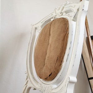 Puede incluir: Silla de madera pintada de blanco con un acabado desgastado. La silla tiene un respaldo alto con un dise&ntilde;o tallado y un coj&iacute;n de asiento redondo y acolchado.