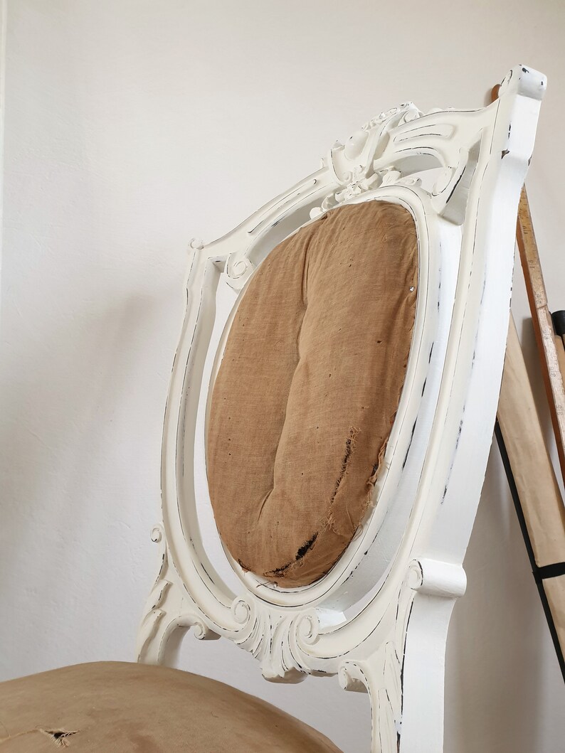 Puede incluir: Silla de madera pintada de blanco con un acabado desgastado. La silla tiene un respaldo alto con un dise&ntilde;o tallado y un coj&iacute;n de asiento redondo y acolchado.