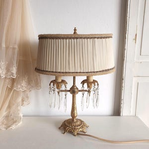 May include: A gold-toned metal table lamp featuring a pleated cream-colored fabric shade with decorative trim. The lamp base has an ornate, textured design and supports two light sockets adorned with hanging clear glass crystal prisms.