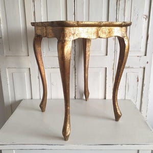 May include: A small, antique gold-colored table with a scalloped edge. The table has four curved legs. The table is approximately 20 inches in diameter and 24 inches tall. The table is made of wood and has a distressed finish.